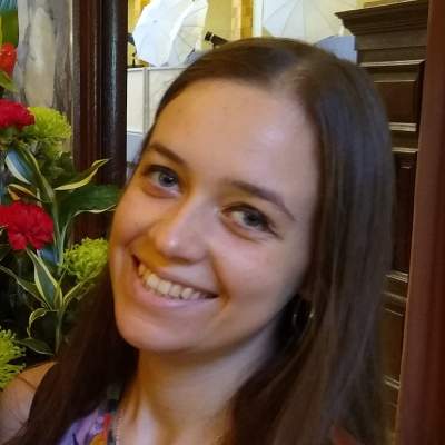 Portrait picture of Maria Czepiel, she is smiling and standing in front of some mahogany panelled wood with flowers in the background. 