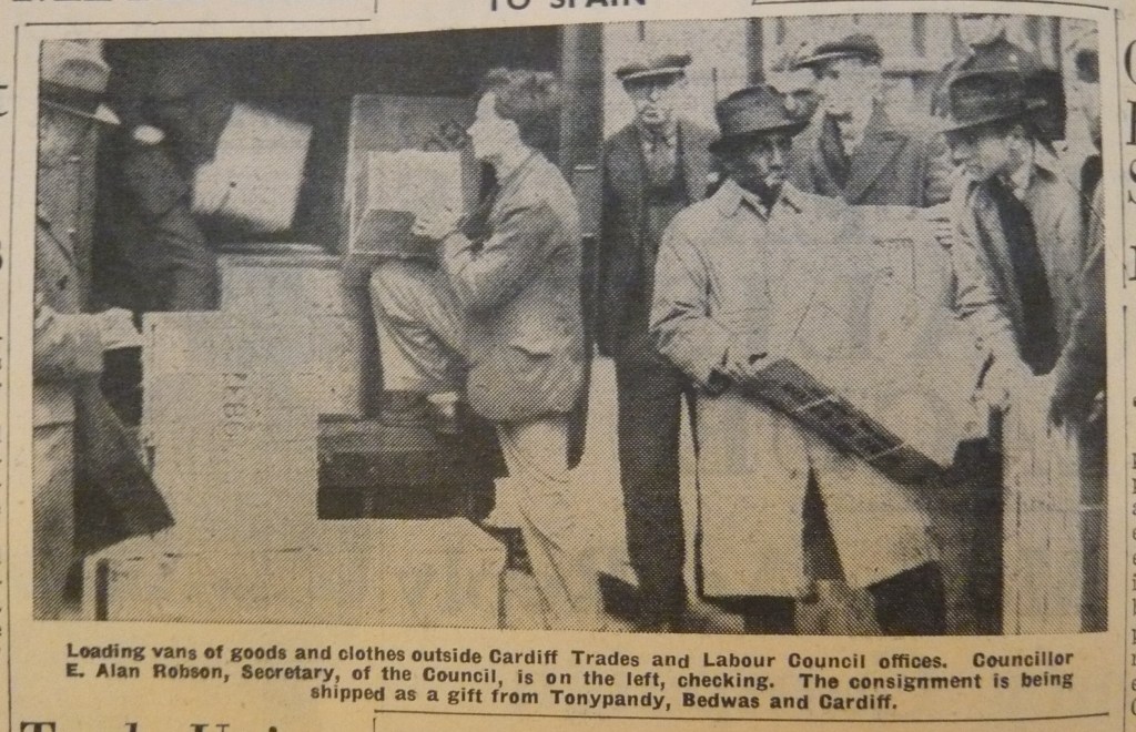 A black-and-white image showing a group of men loading goods and clothes into vans outside the Cardiff Trades and Labour Council offices. One man, identified as E. Alan Robson, the Secretary of the Council, is on the left checking the consignment. The other individuals are assisting in carrying large packages.