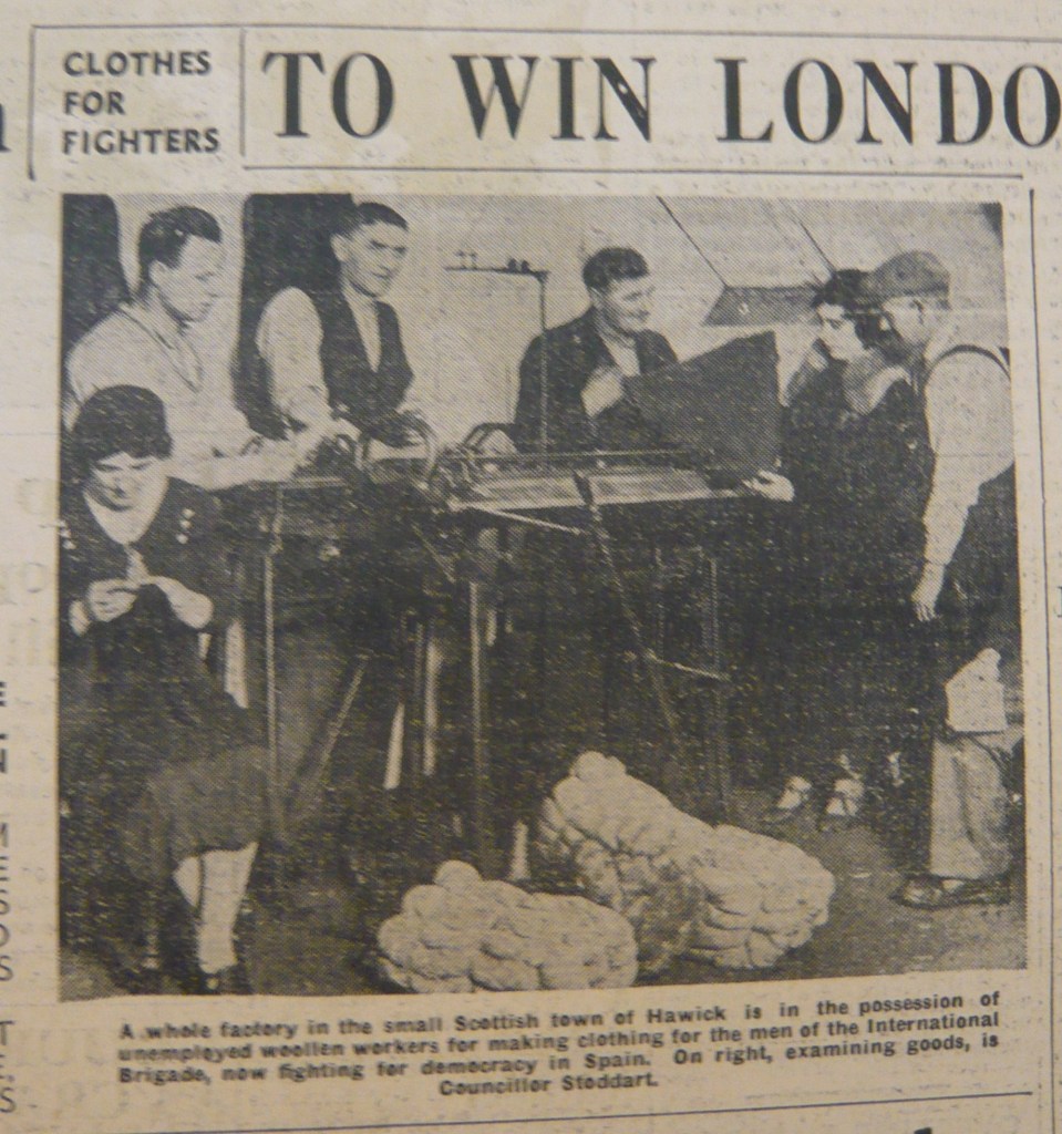 A black-and-white image showing a group of people working in a factory in Hawick, Scotland. They are seated at tables knitting and inspecting woolen goods. On the right, Councillor Stoddart is shown examining finished items. The image features large bundles of wool in the foreground.