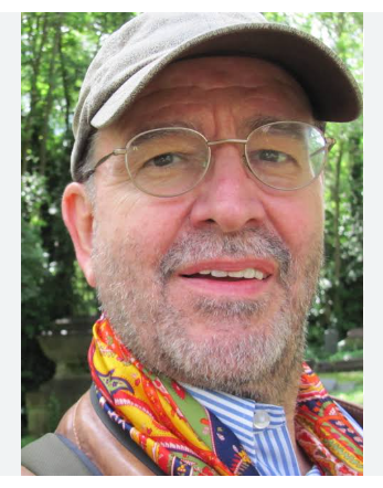 A picture of Professor Benjamin Davy, he is wearing a light grey cap, wire rimmed glasses, a blue and white striped shirt, and a colourful scarf. He is looking in to the camera with a half smile.