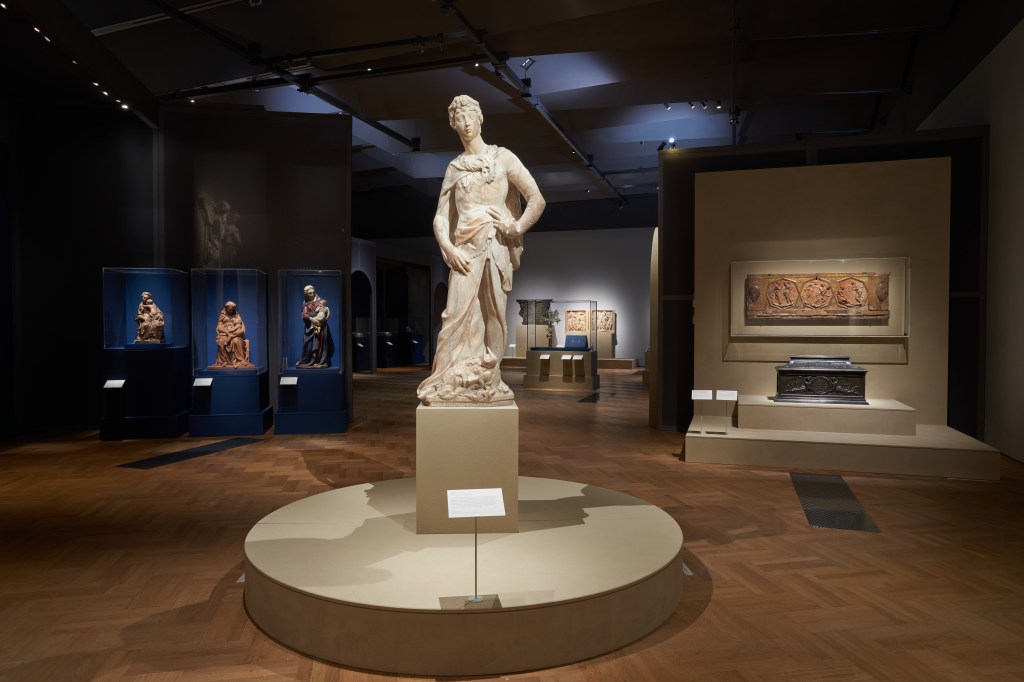 View of the exhibition Donatello: Sculpting the Renaissance, 2023, Victoria and Albert Museum, London (photo: Peter Kelleher © Victoria and Albert Museum, London). 
The photograph shows a sculpture by Donatello sitting on a plinth in a softly lit exhibition space. The sculpture is a pale stone or marble and depicts a man standing in a classical pose wearing a long cloth which is gathered at his feet.