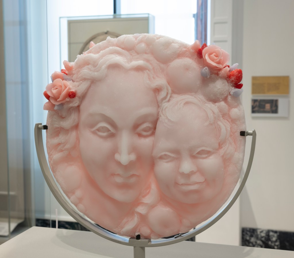 Rebecca Stevenson, A Tray of Sweetmeats, 2023, wax and resin, 27 x 27 x 6 cm.  London, Rebecca Stevenson (photo: Peter Kelleher © Victoria and Albert Museum, London). The art work is a pale pink disc suspended on a metal stand. The disc depicts a the face of a woman and a child, their faces close together, darker pink roses with touches of red sit above their heads.