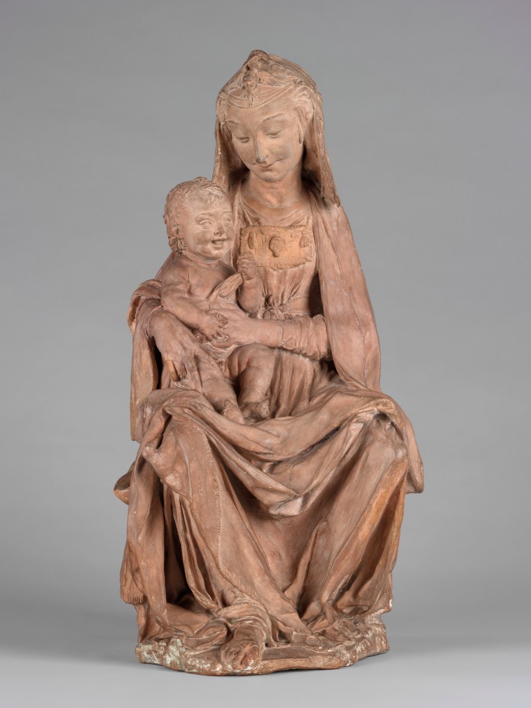 The seated Virgin gazes sweetly down at her laughing baby, his mouth open and tongue visible. She holds him in her right arm, as he plays with his swaddling, her long fingers splayed on his right thigh. A jewelled clasp holds the folds in her veil above her oval face; a decorative panel hangs across her chest. Her cloak and gown fall in generous folds around her lap.