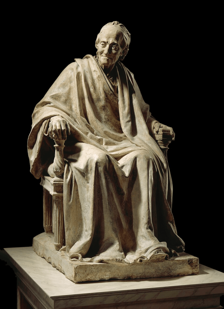 A white marble sculpture depicting a seated Voltaire wrapped in cloth. 